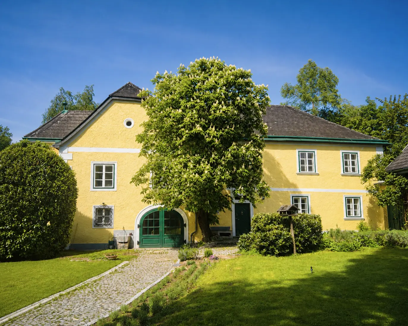 A bright yellow two-story house with a dark roof, green arched doors, and a large flowering tree in a lush green garden.