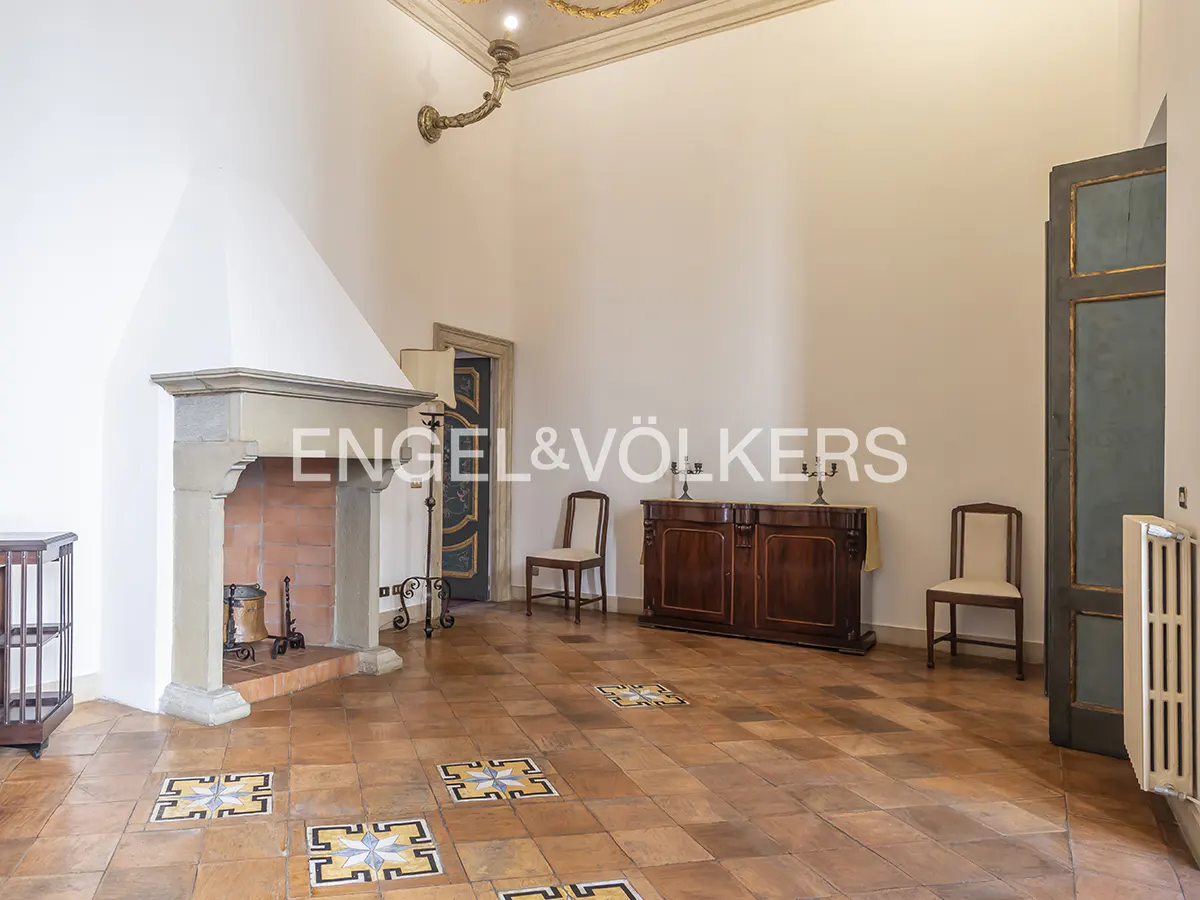 A spacious room with a stone fireplace, terracotta tile floor, and antique furniture. The walls are white, and the ceiling has decorative molding.