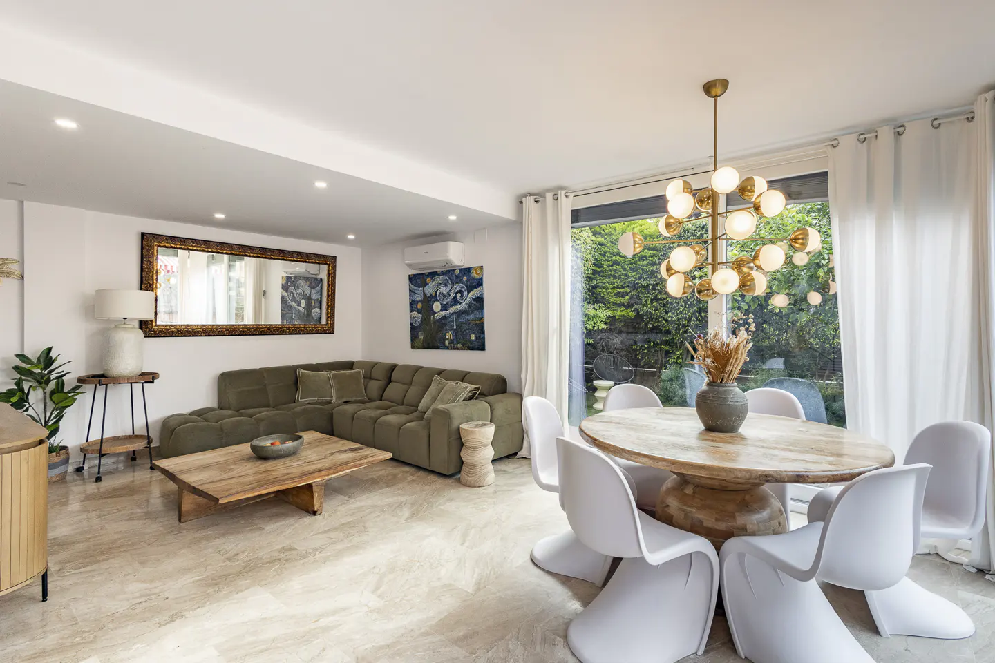 Bright living and dining area with green sectional sofa, wood table, and white chairs. A modern chandelier hangs above the dining table.