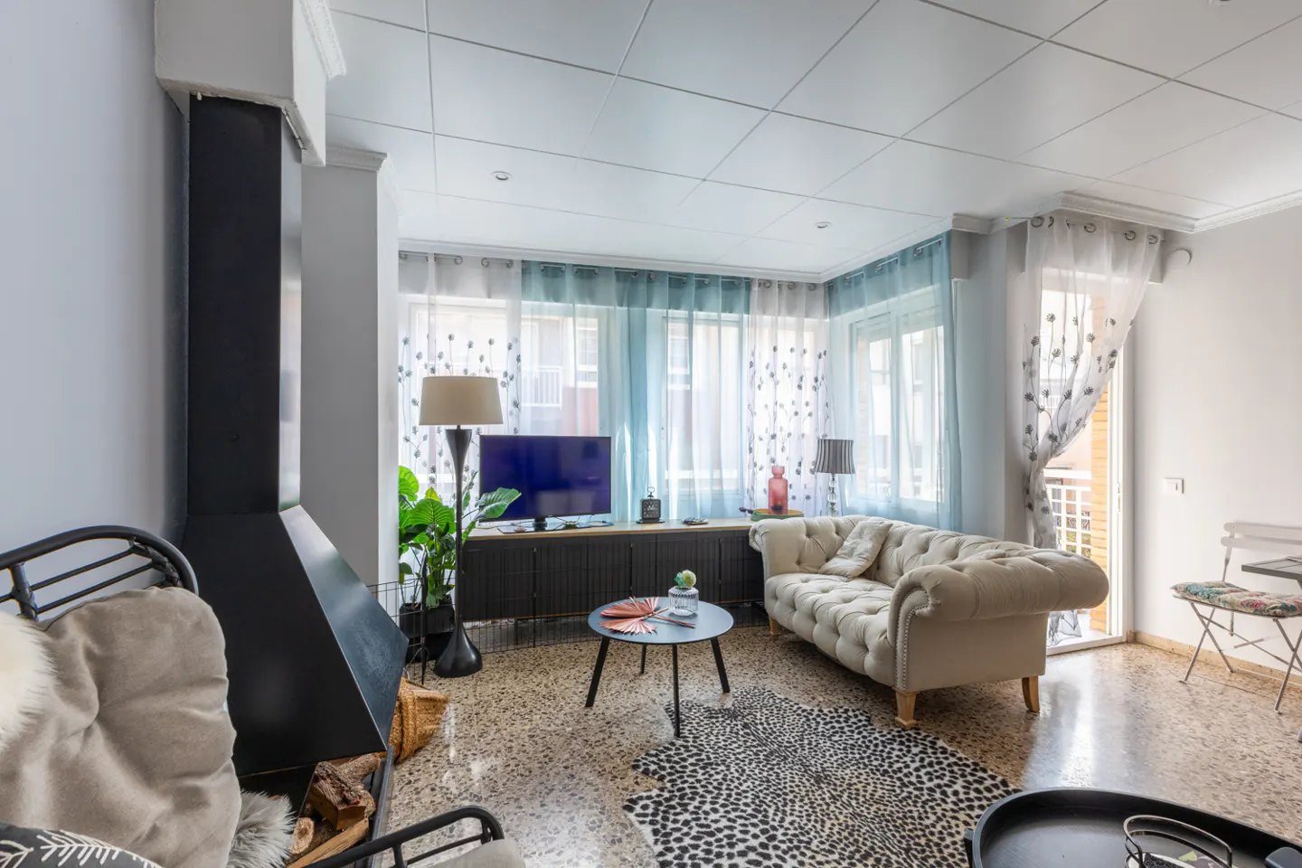 Living room with a black fireplace, beige sofa, TV, and a black and white animal print rug. The walls are white and the curtains are blue and white.