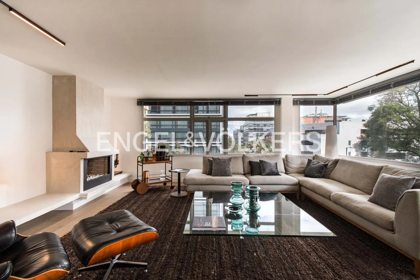 Living room with a gray sectional sofa, glass coffee table, fireplace, and large windows with city views. Black leather chair and bar cart.