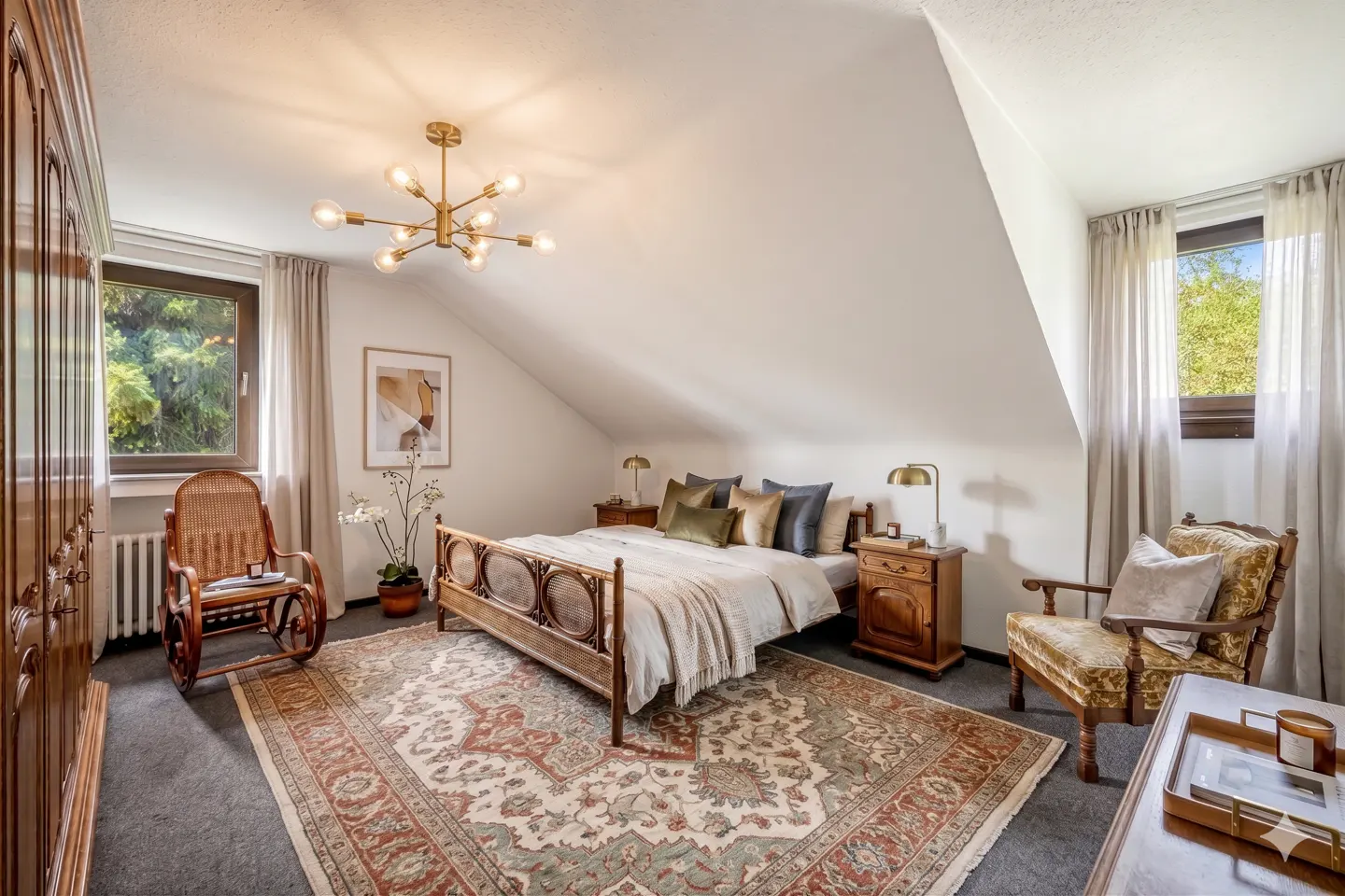 Attic bedroom with a wooden bed, nightstands, and a patterned rug. A gold chandelier hangs from the white ceiling. Windows with sheer curtains.