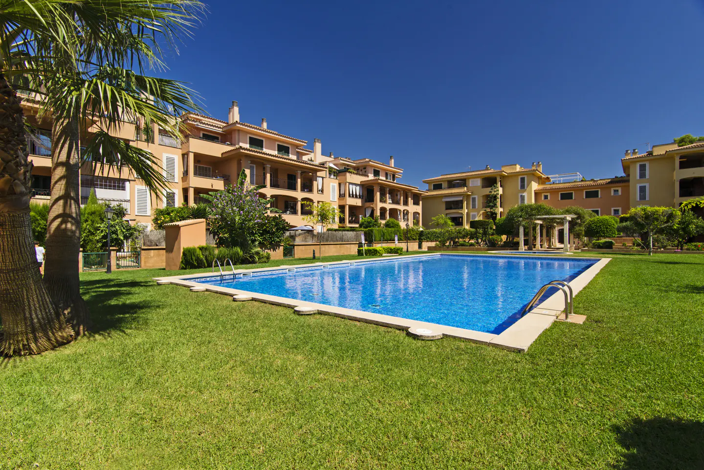 A bright blue pool is surrounded by green grass and peach-colored buildings under a clear blue sky.