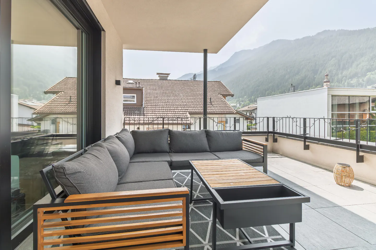 Balcony with gray sectional sofa, wood table, and mountain view. Black and white patterned rug. Outdoor seating area.