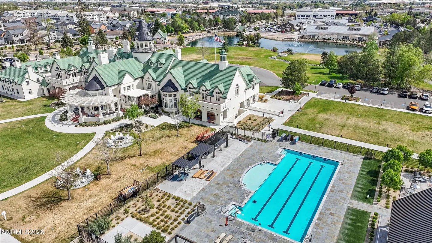 Aerial view of a white clubhouse with green roofs, a pool, and a pond in a suburban area.