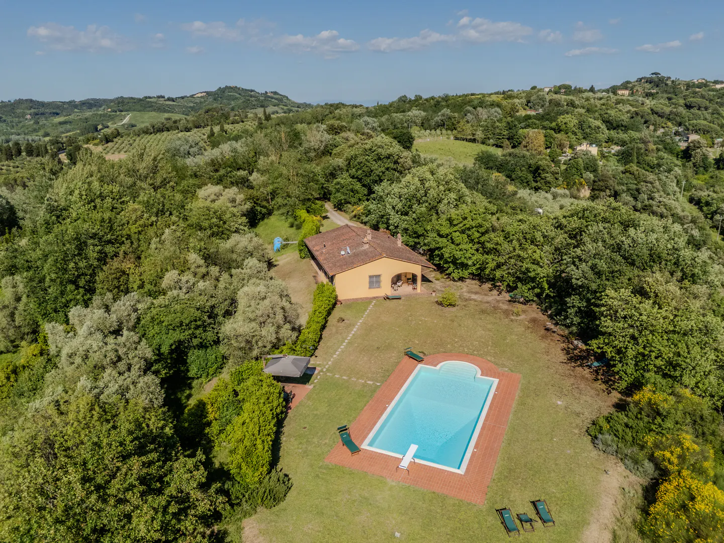 Aerial view of a yellow house with a red tile roof, surrounded by green trees, and a rectangular pool with blue water.