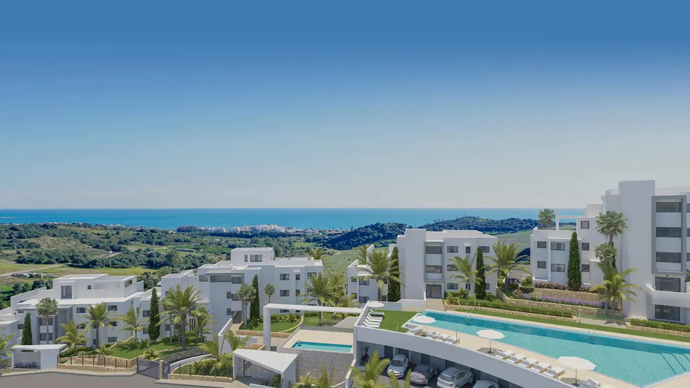 Aerial view of modern white apartments with pools, palm trees, and ocean backdrop under a clear blue sky.