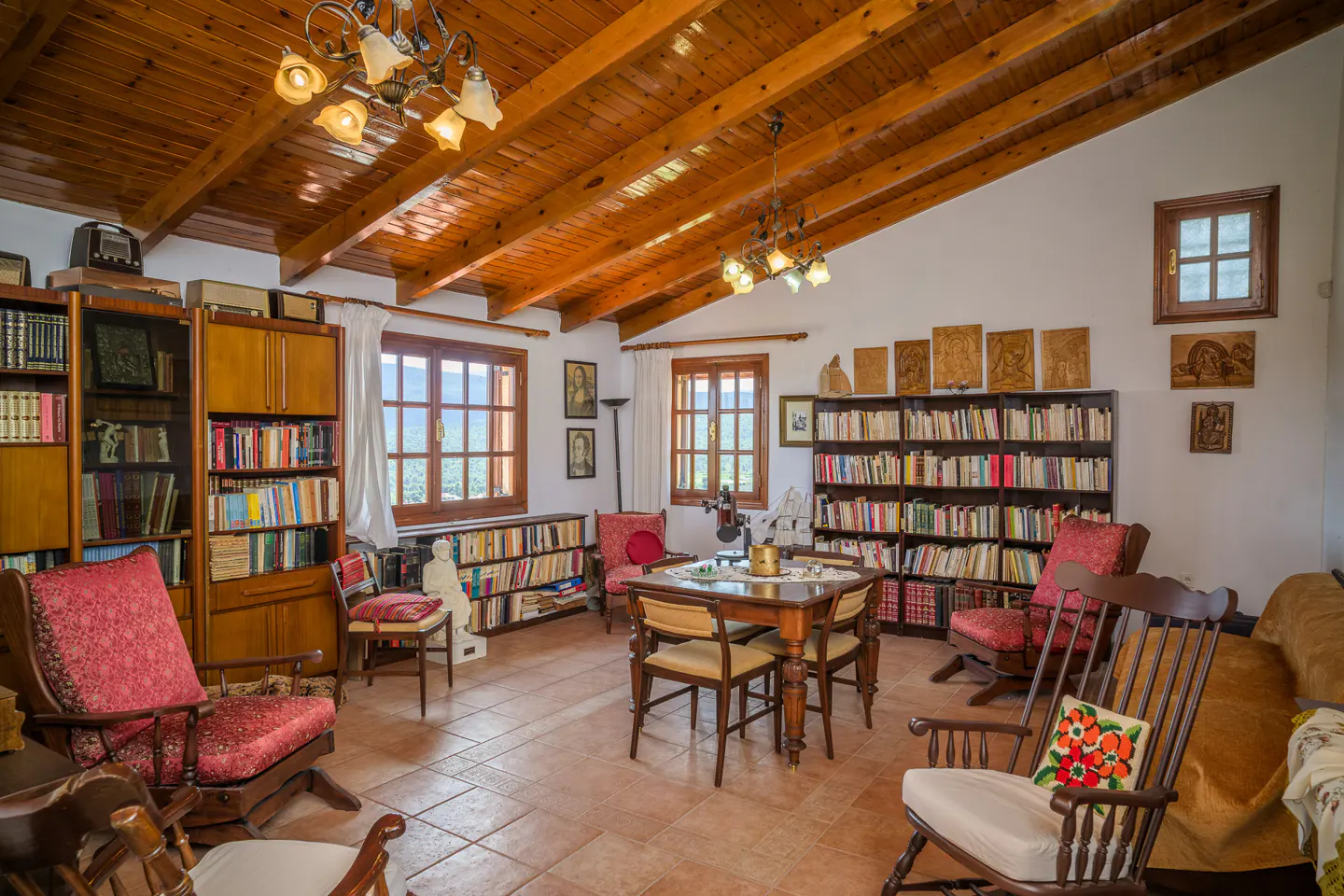 A cozy living room with wooden ceiling, bookshelves, chairs, and a table. Natural light streams through the windows.