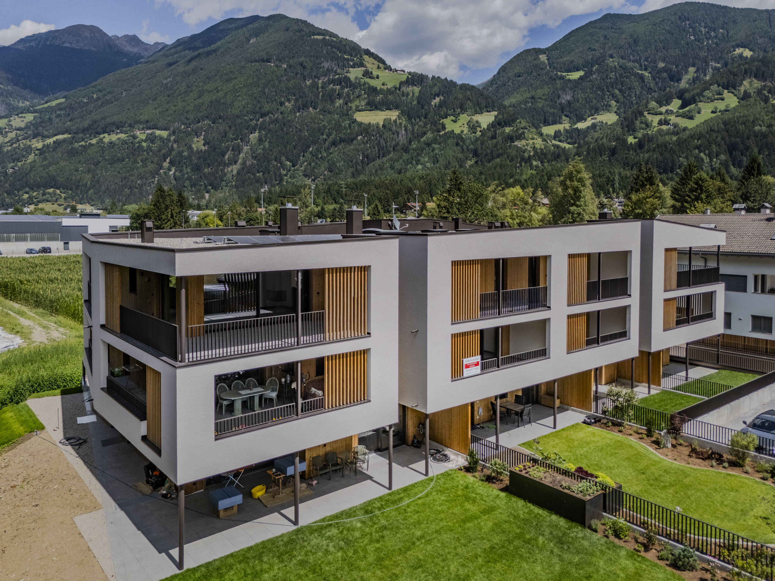 Modern multi-story apartment building with balconies and wooden accents, set against a backdrop of lush green mountains.