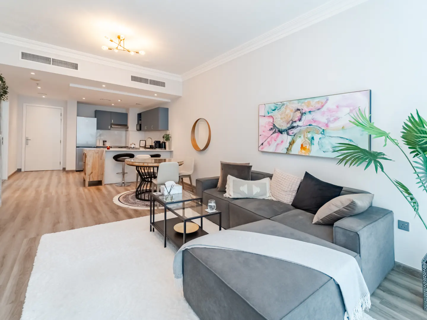 Open-concept living space with gray sectional, glass coffee table, and abstract art. Kitchen with gray cabinets and island in background.