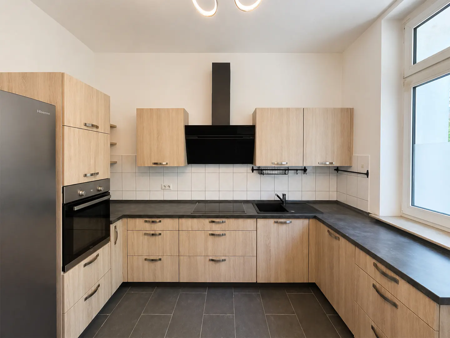 A modern kitchen with light wood cabinets, black countertops, and a stainless steel refrigerator.