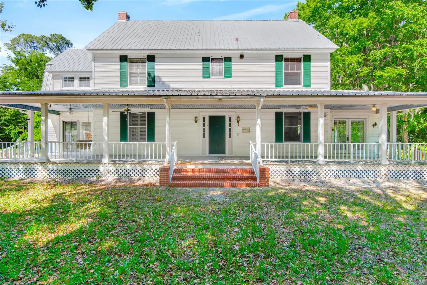 Two-story white house with green shutters and a metal roof. A wide porch with white railings wraps around the front.