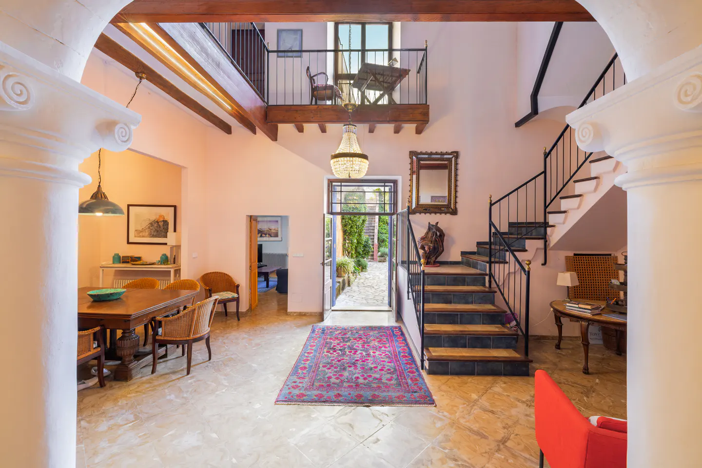 Interior view of a home with pink walls, tile floors, and a staircase. A balcony overlooks the entrance, which leads to a garden.