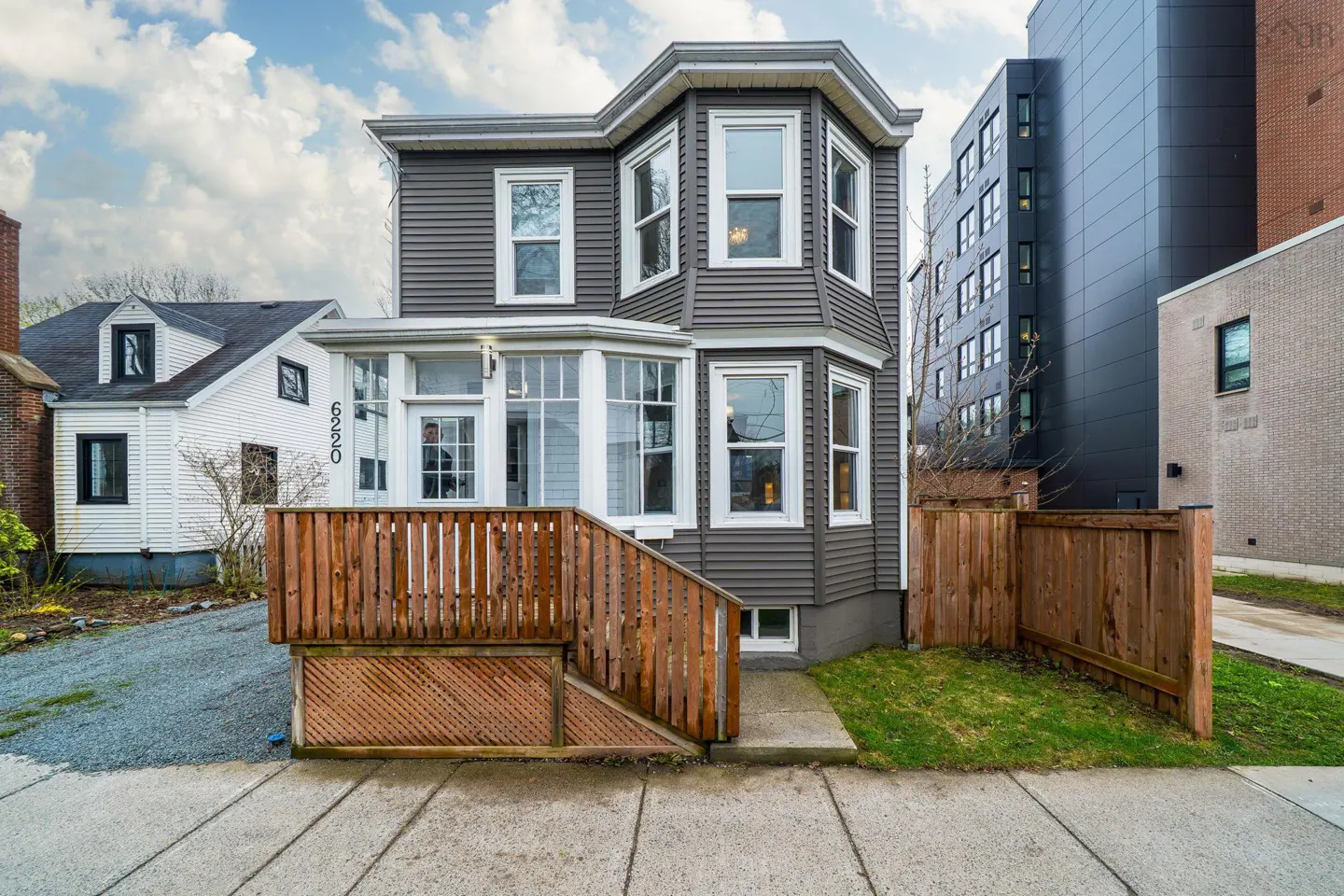Two-story gray house with white trim, a wooden porch, and a fenced yard. A modern building is in the background.