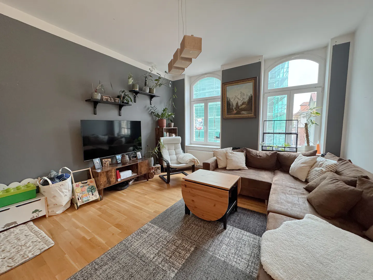 Living room with gray walls, wood floors, brown sectional, and large windows. Shelves with plants are above the TV. A light wood table sits on a gray patterned rug.