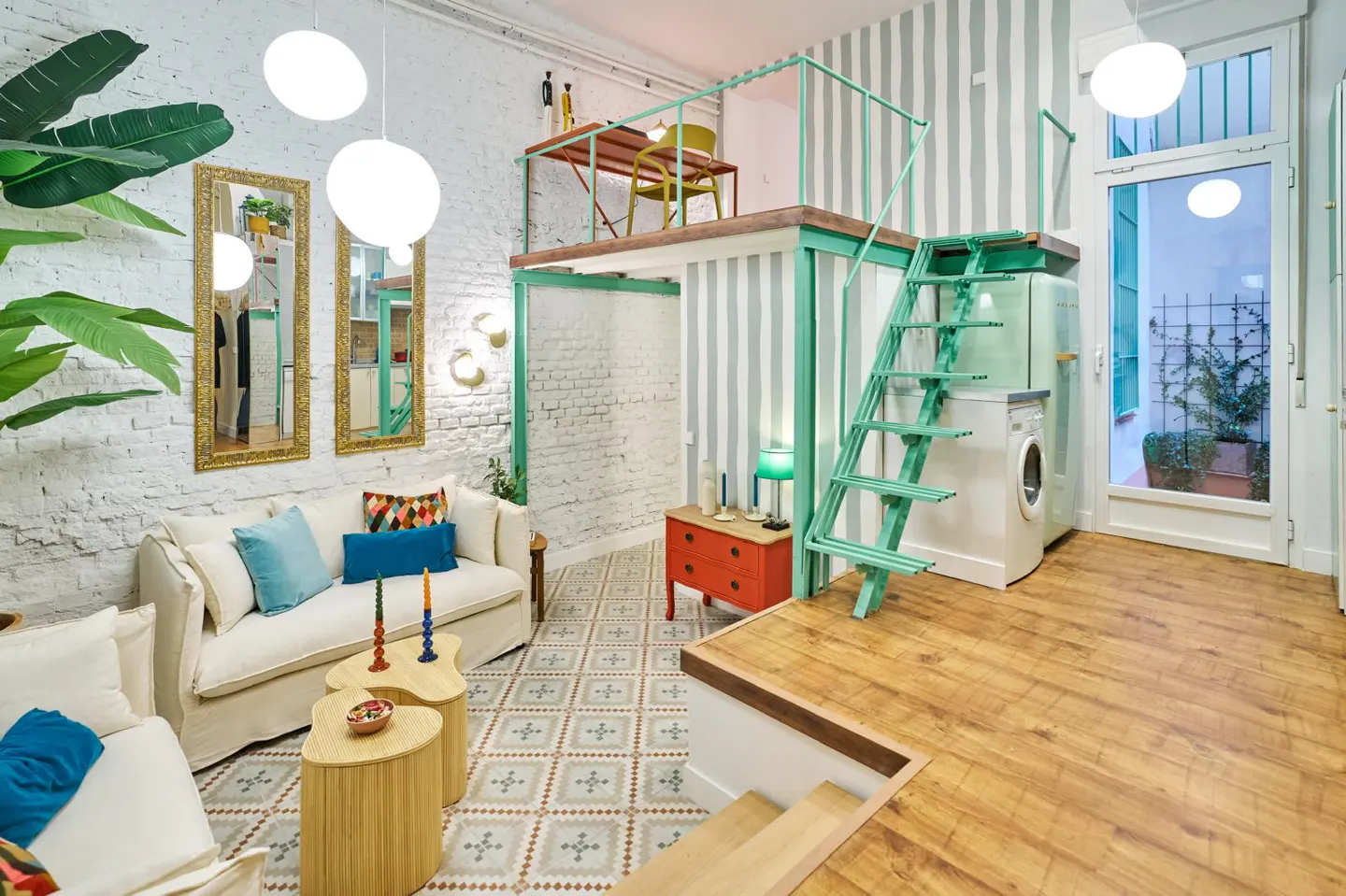 Bright, modern apartment interior with white sofas, patterned tile, and a loft with green railings and stairs. A washing machine sits near a glass door.