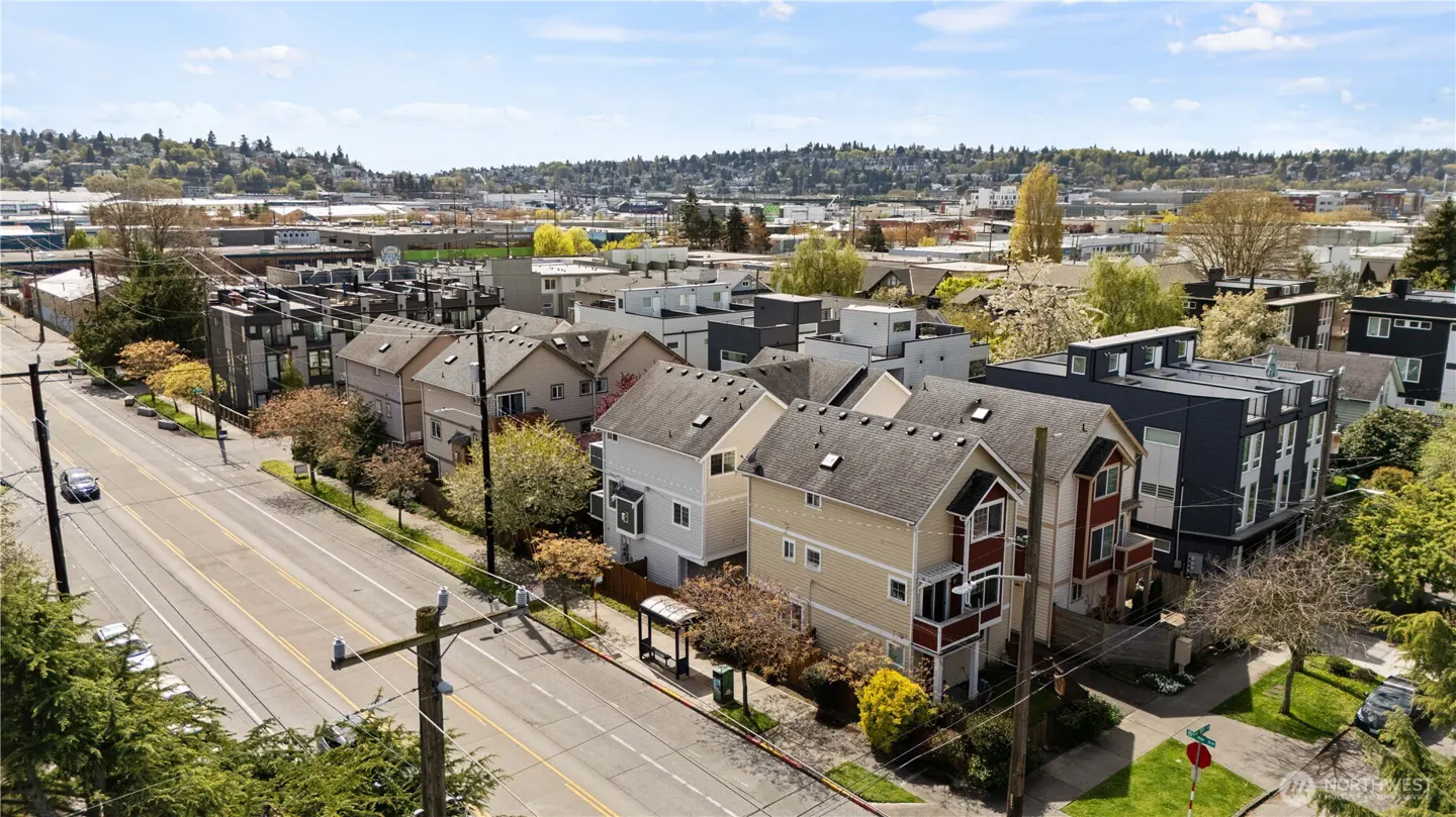 Aerial view of a street with modern townhouses in varying colors, trees, and a city skyline under a blue sky.