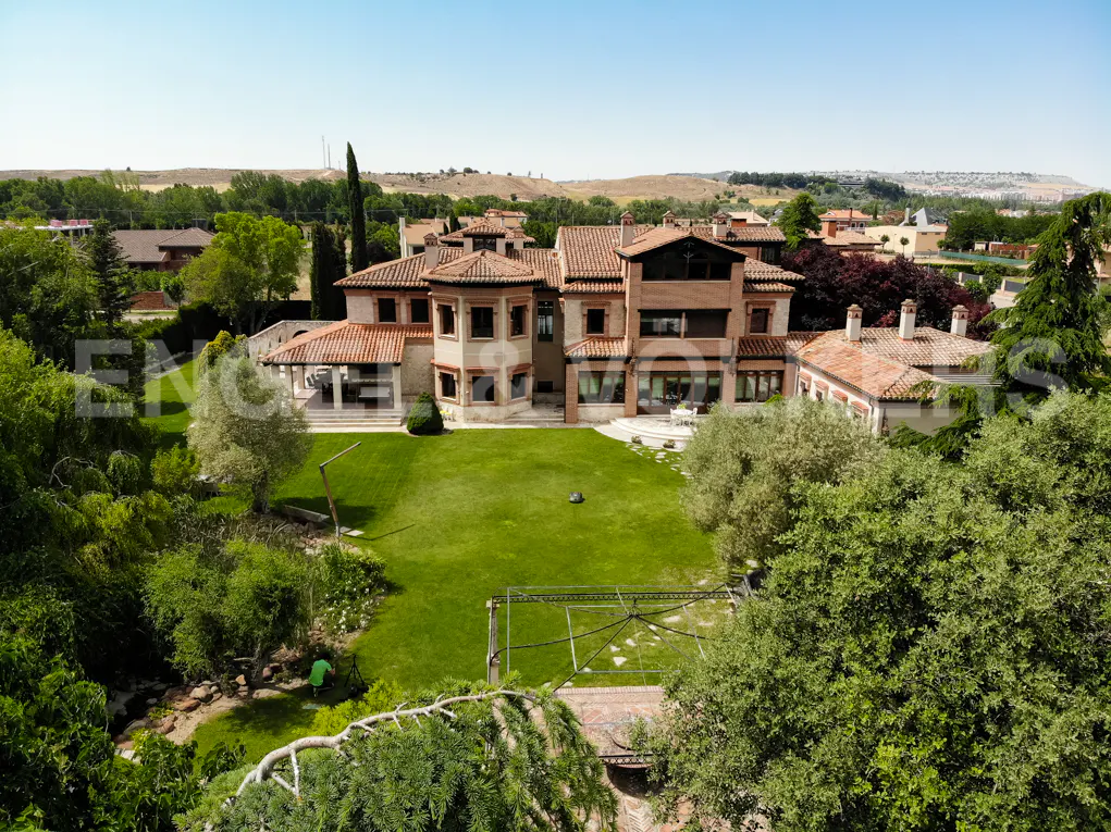 Aerial view of a large Mediterranean-style villa with a terracotta roof, surrounded by a lush green lawn and mature trees.
