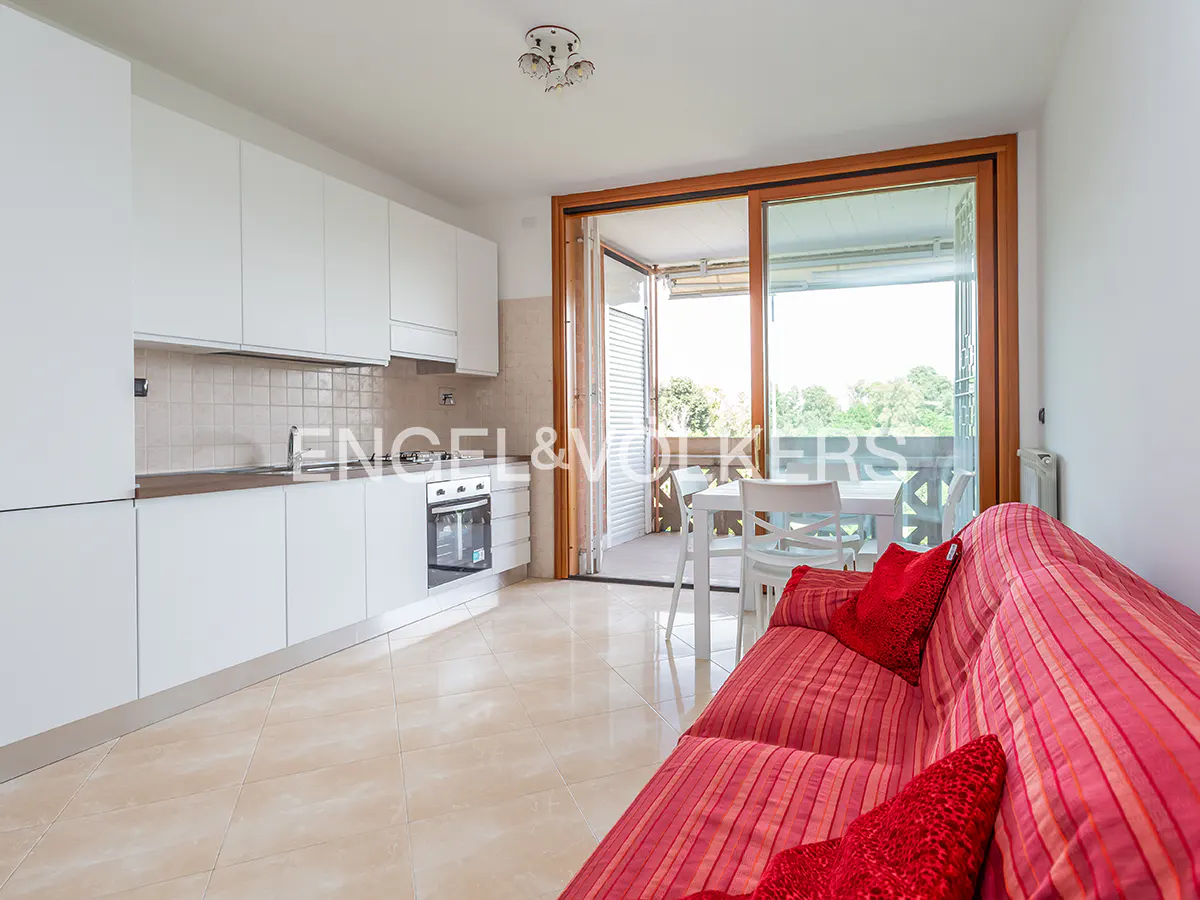 Bright apartment interior with white kitchen cabinets, red striped sofa, and balcony with table and chairs.