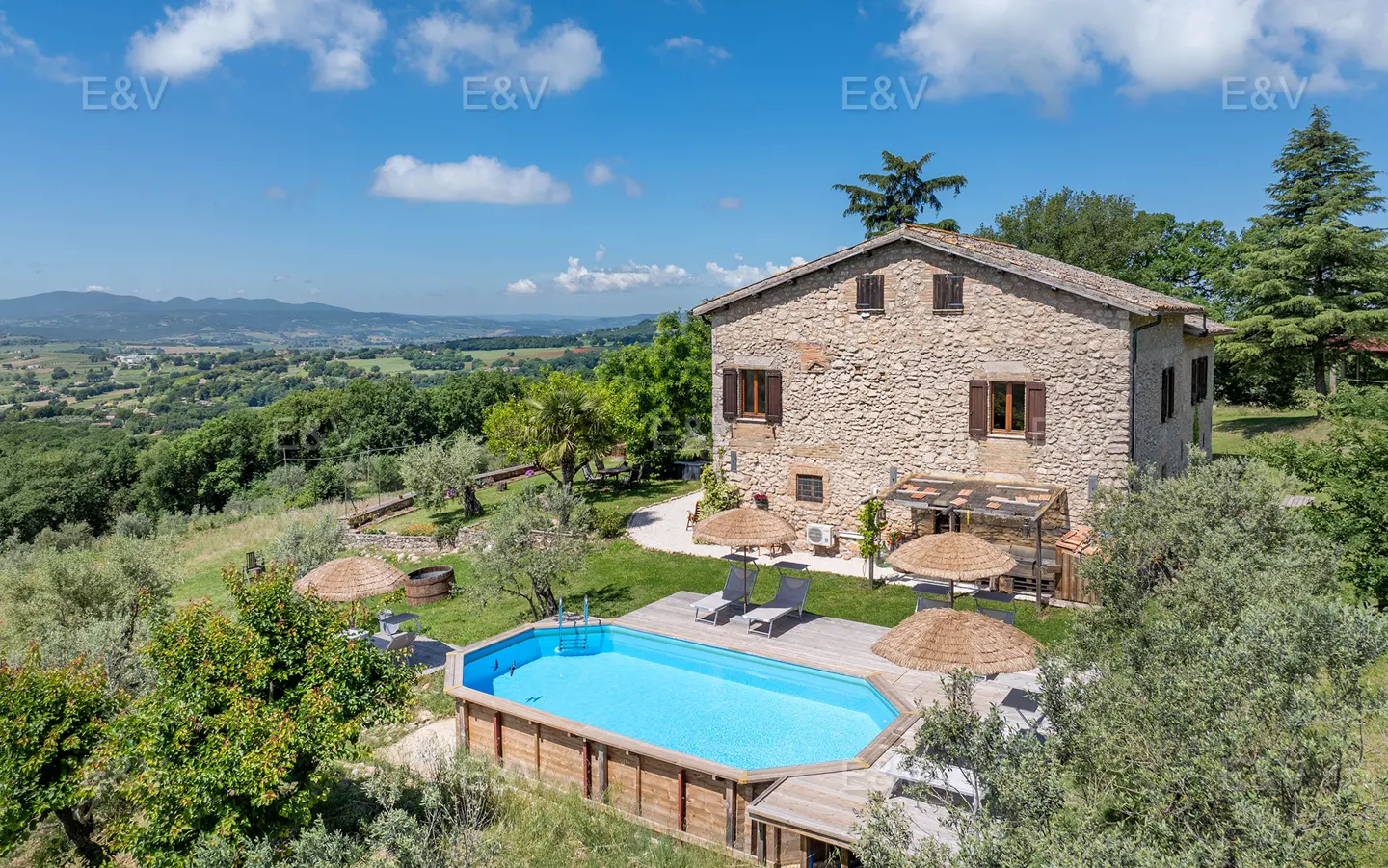 A stone farmhouse with a pool, wooden deck, and straw umbrellas overlooking a lush green valley under a blue sky.
