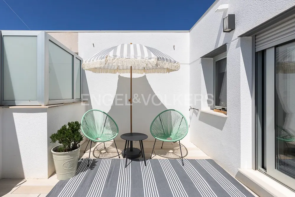 A sunny patio with two green chairs, a striped umbrella, and a small table on a striped rug. The walls are white.