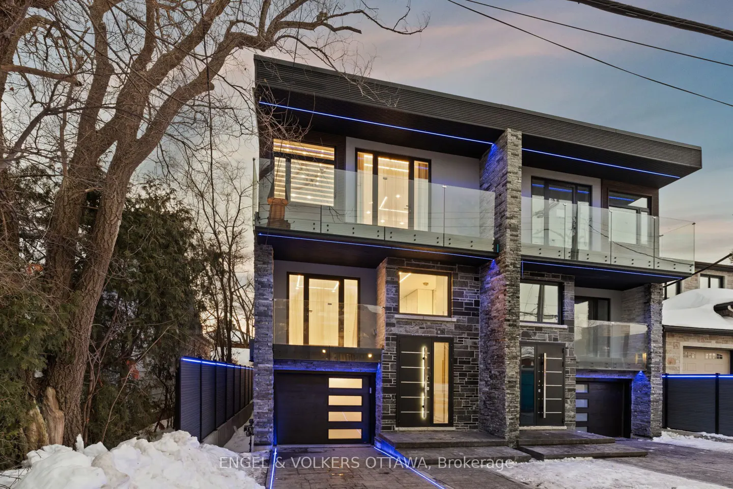 Modern two-story duplex with stone accents, black trim, glass balconies, and blue LED lighting at dusk.