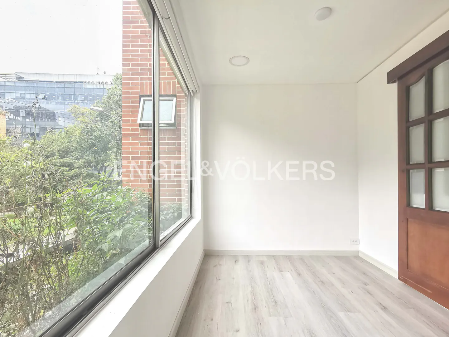 Bright, empty room with light wood floors, white walls, and a large window overlooking greenery and a brick building. A wooden door is on the right.