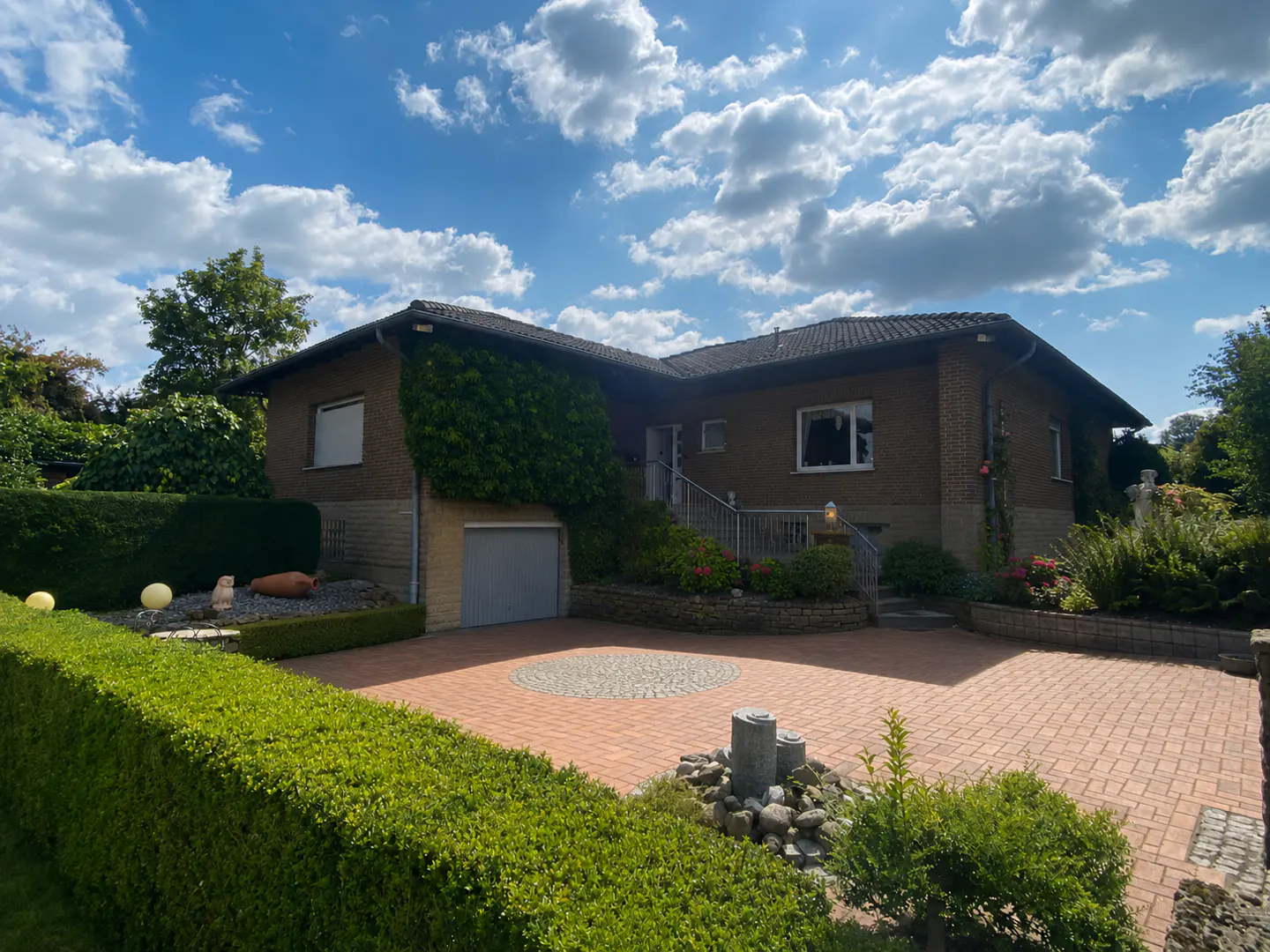 A single-story brick house with green ivy, a gray garage door, and a brick driveway under a blue sky.