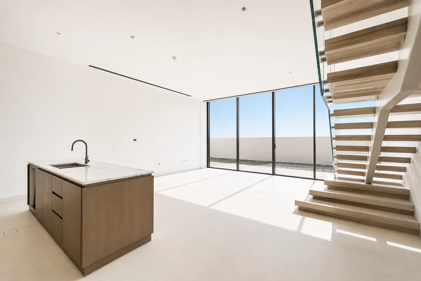 Bright, modern interior with white walls, light floor, and wood stairs with glass railing. A kitchen island with a marble top stands near a large window.