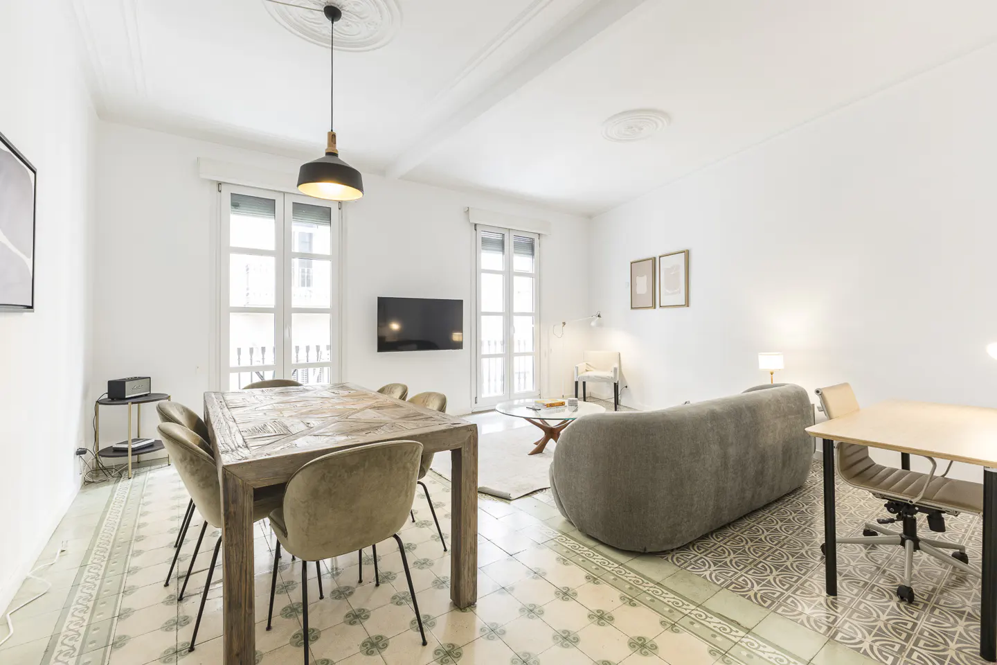 Bright, open-concept living space with a dining table, sofa, and desk. White walls and patterned tile flooring. Natural light from windows.