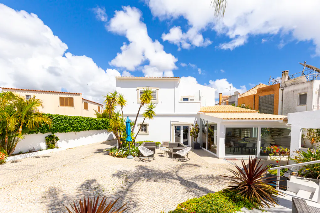 Backyard of a white house with a stone patio, outdoor furniture, palm trees, and a glass-enclosed sunroom under a blue sky.
