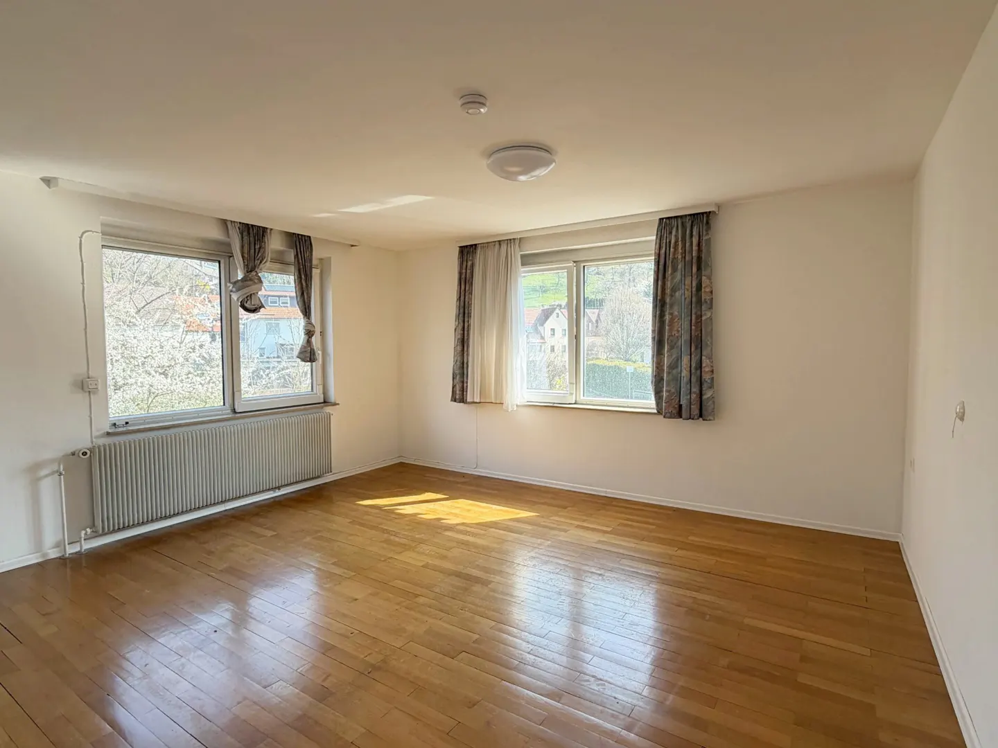 Bright, empty room with hardwood floors, white walls, and two windows with patterned curtains. A radiator sits under one window.