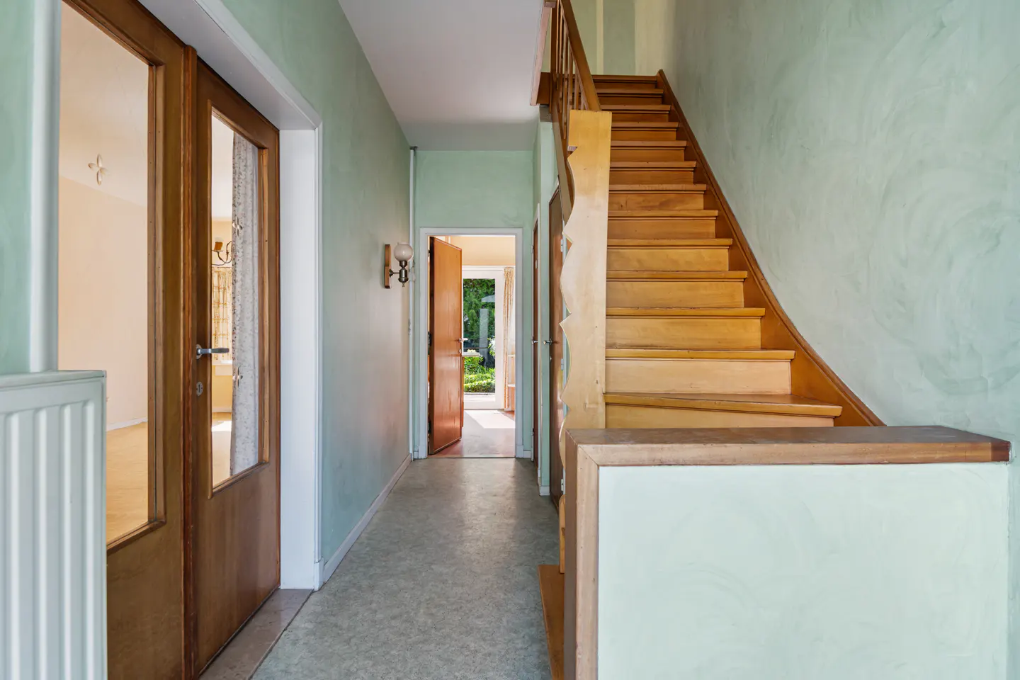 Hallway with light green walls, wooden staircase, and open door leading to a garden view. A glass-paneled door is on the left.