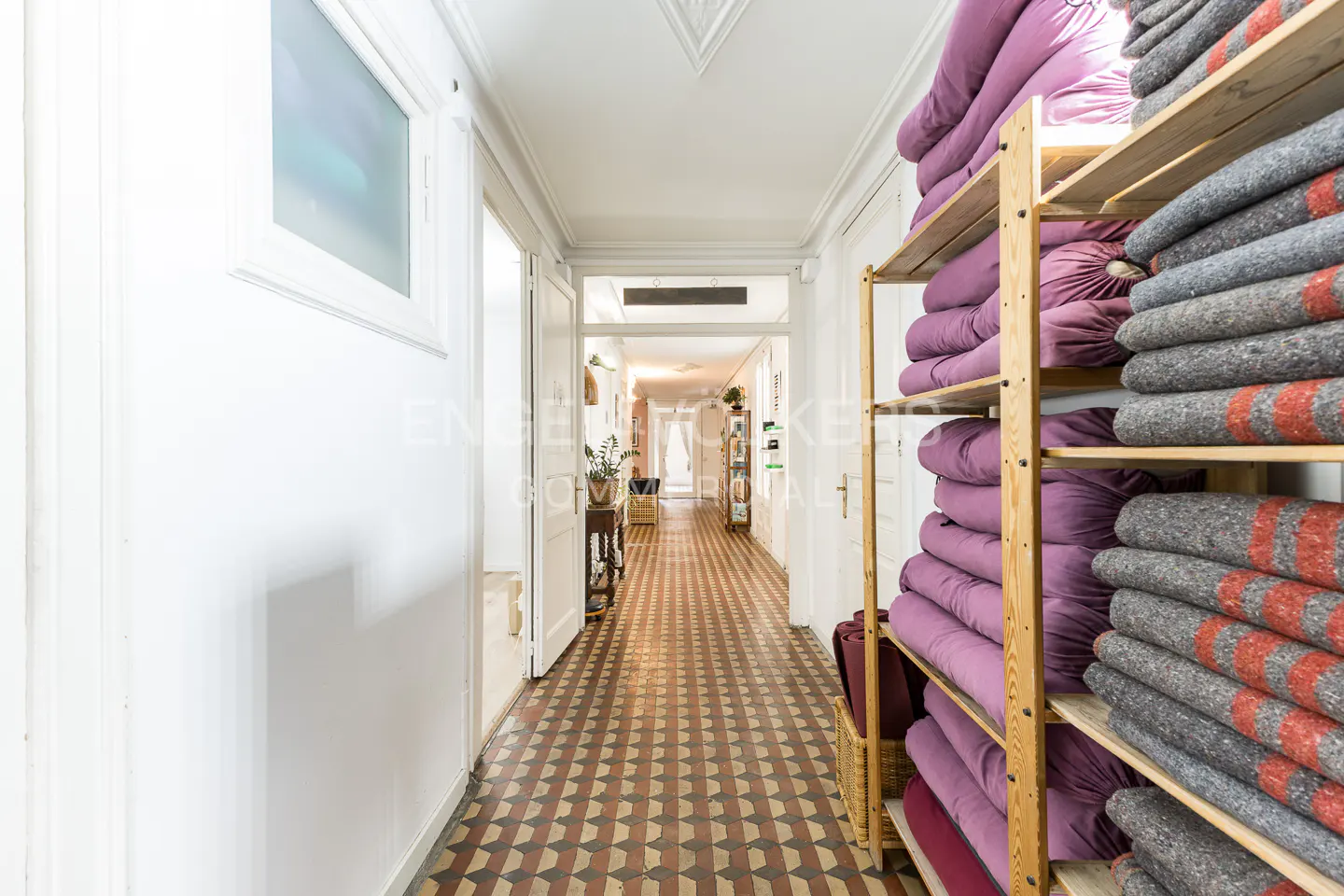 Hallway view with patterned tile floor, white walls, and wooden shelves stacked with purple cushions and gray blankets.