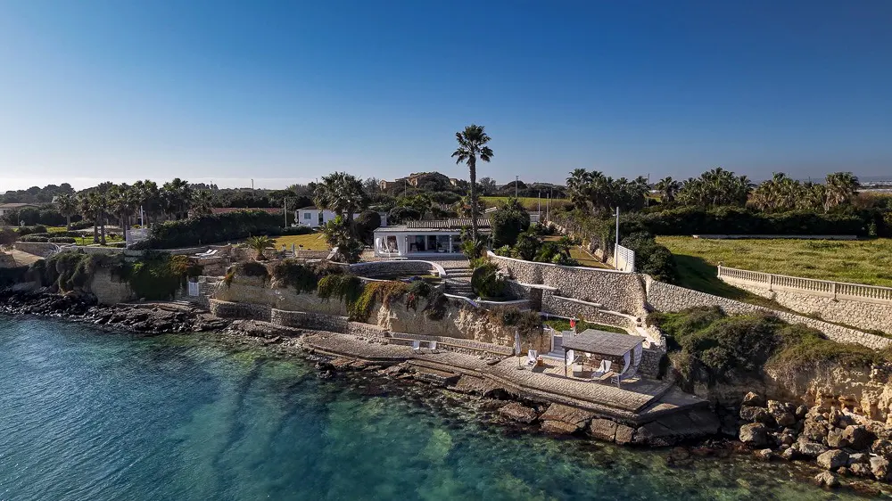 Oceanfront property with a white house, palm trees, and stone walls on a cliff overlooking turquoise water under a clear blue sky.