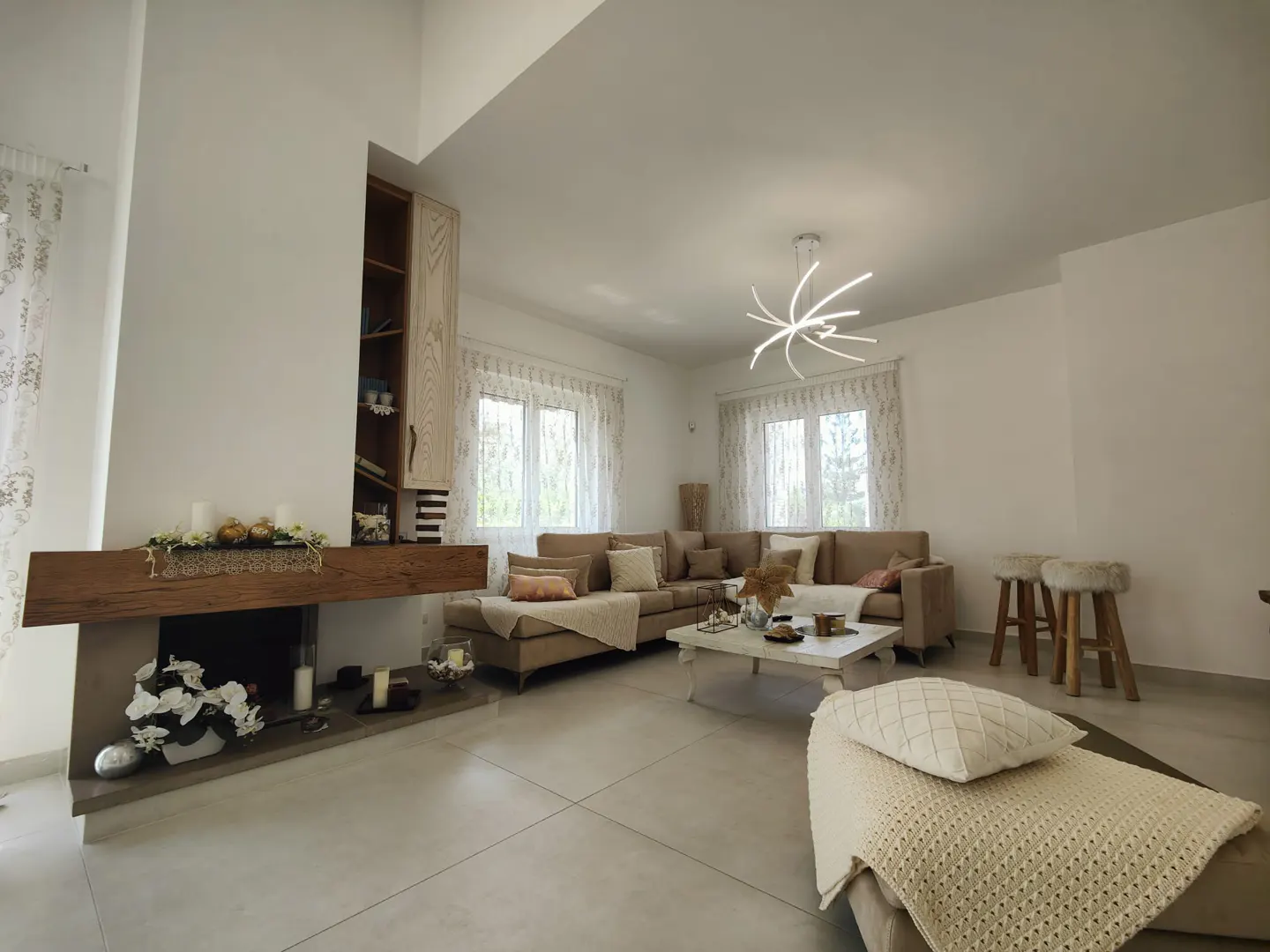 Bright living room with a beige sectional sofa, white walls, and a modern light fixture. A fireplace with a wooden mantel and built-in shelves adds character.