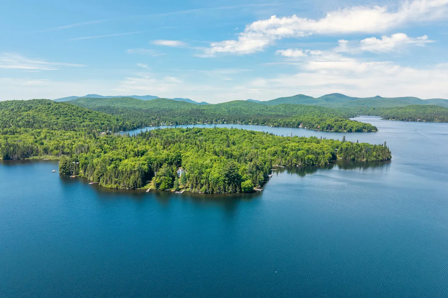 Aerial view of a lush green forested peninsula extending into a deep blue lake under a bright, partly cloudy sky.