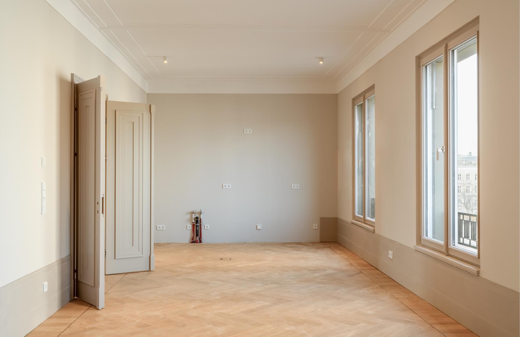 Empty room with light beige walls, herringbone wood floors, two tall windows, and open paneled doors.