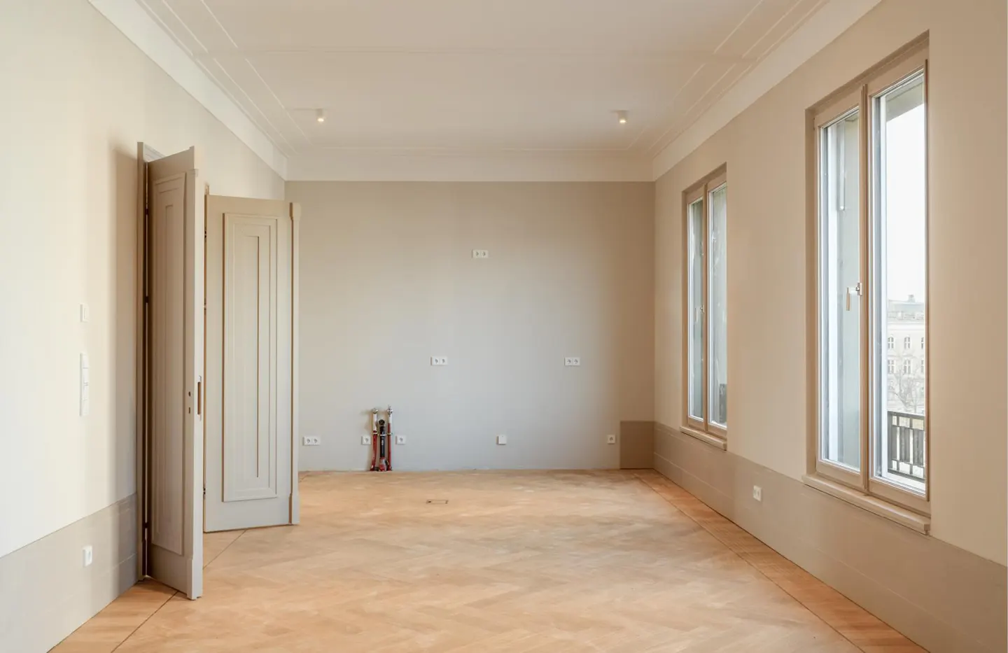 Empty room with light beige walls, herringbone wood floors, two tall windows, and open paneled doors.