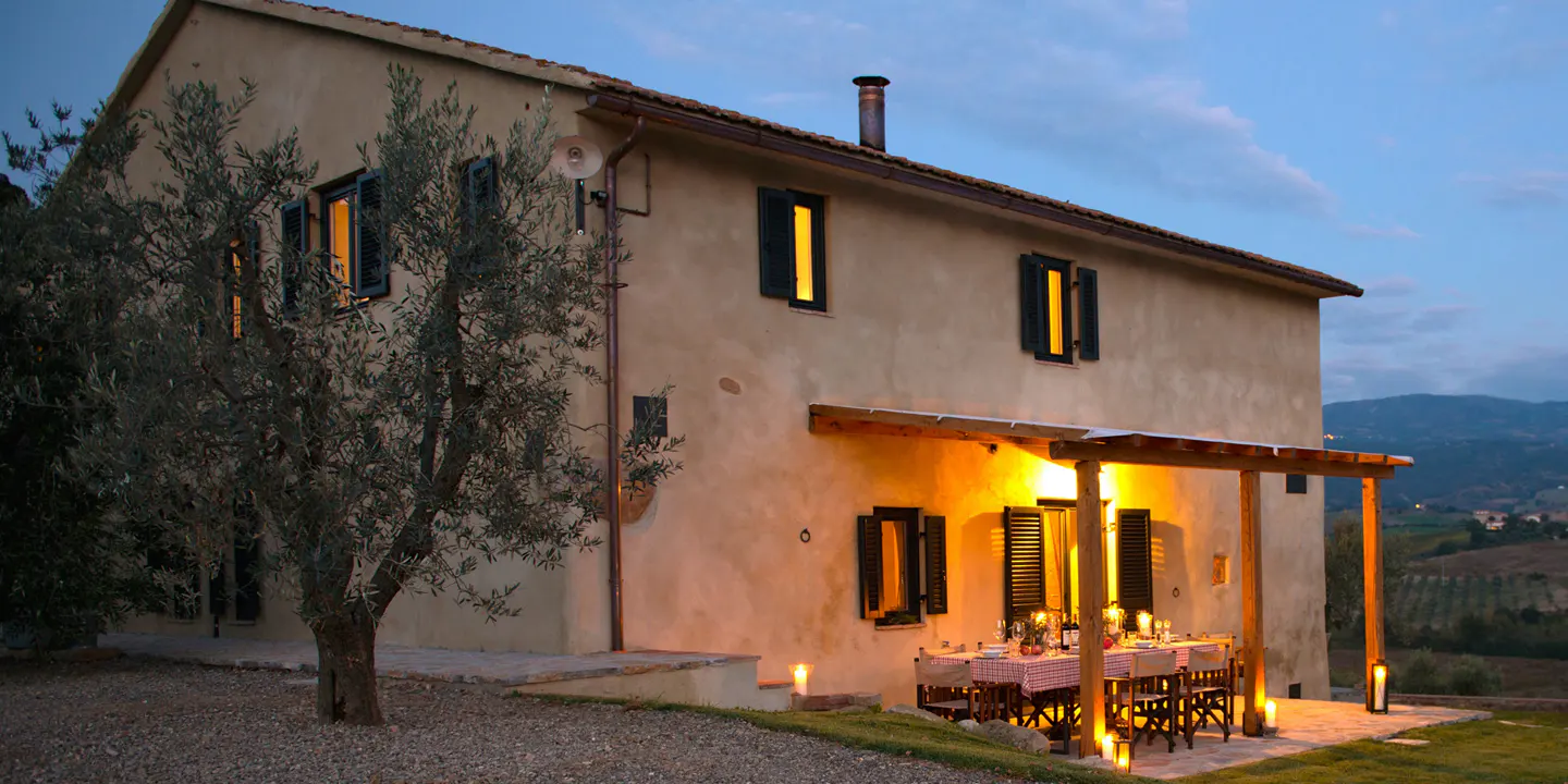 Exterior view of a two-story beige house with dark shutters and a covered patio with a set table at dusk.