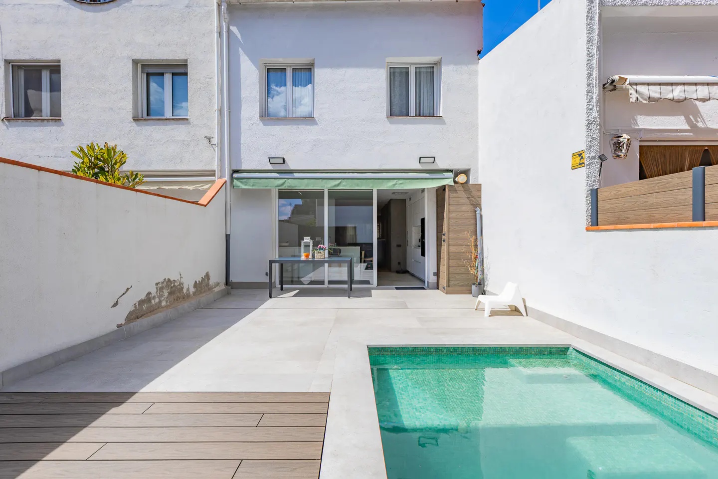 A backyard with a small pool, a table with drinks, and a white plastic chair. The house is white with a green awning.