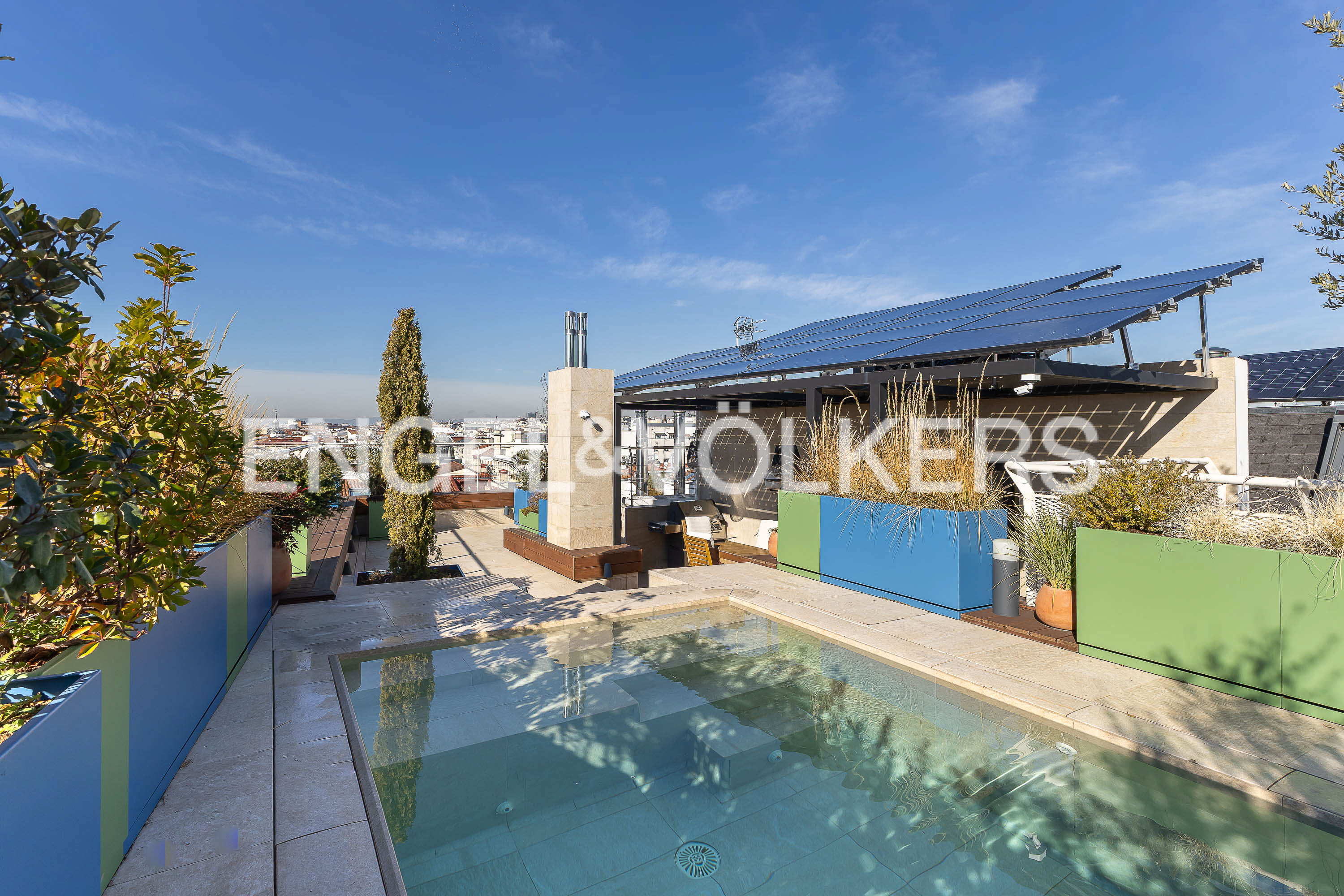 Rooftop terrace with a pool, blue and green planters, and solar panels under a blue sky.