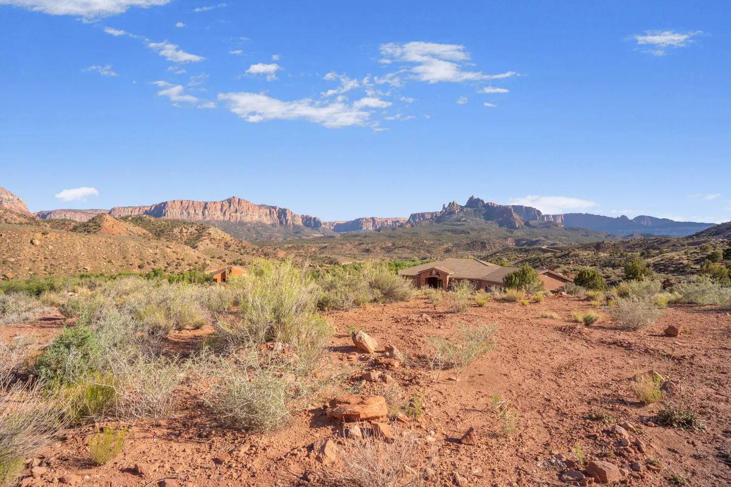 Desert landscape with a house, red dirt, and mountains under a blue sky with scattered clouds.