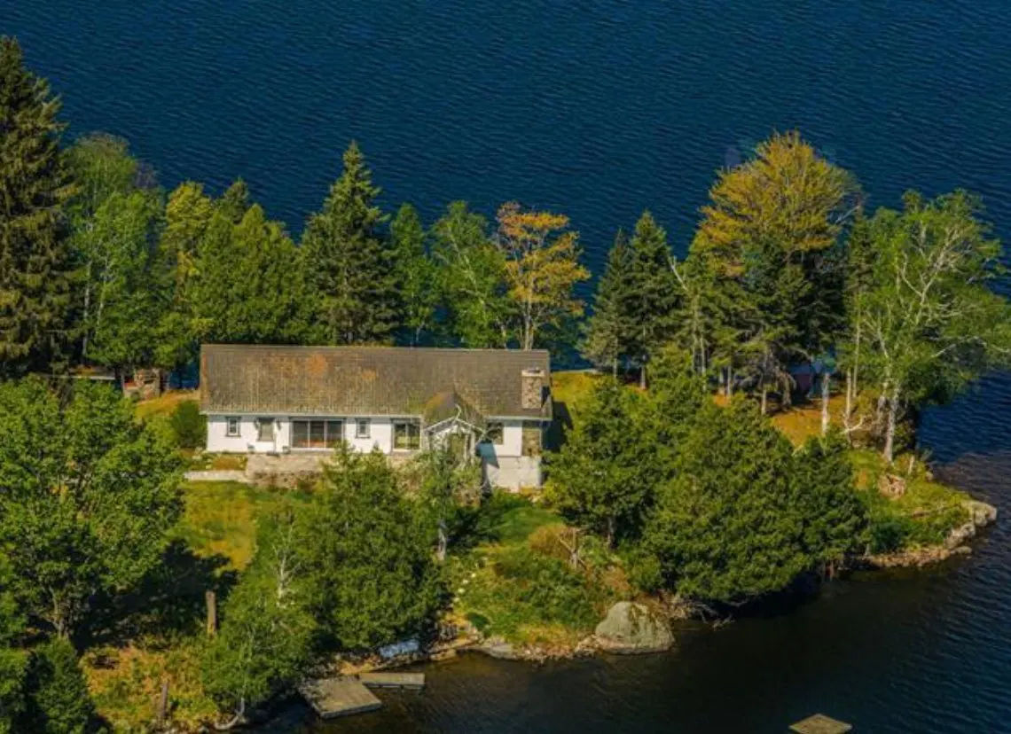 Aerial view of a white house with a brown roof, surrounded by green trees on a small island in a blue lake.