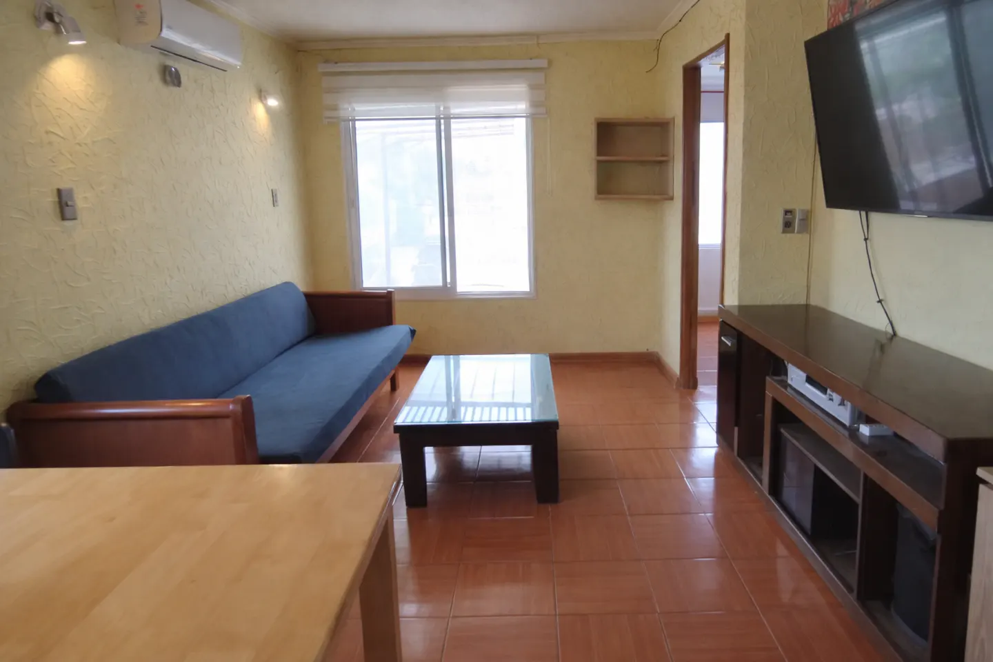 A living room with yellow walls, brown tile floors, a blue sofa, a glass coffee table, and a TV on a dark wood stand.