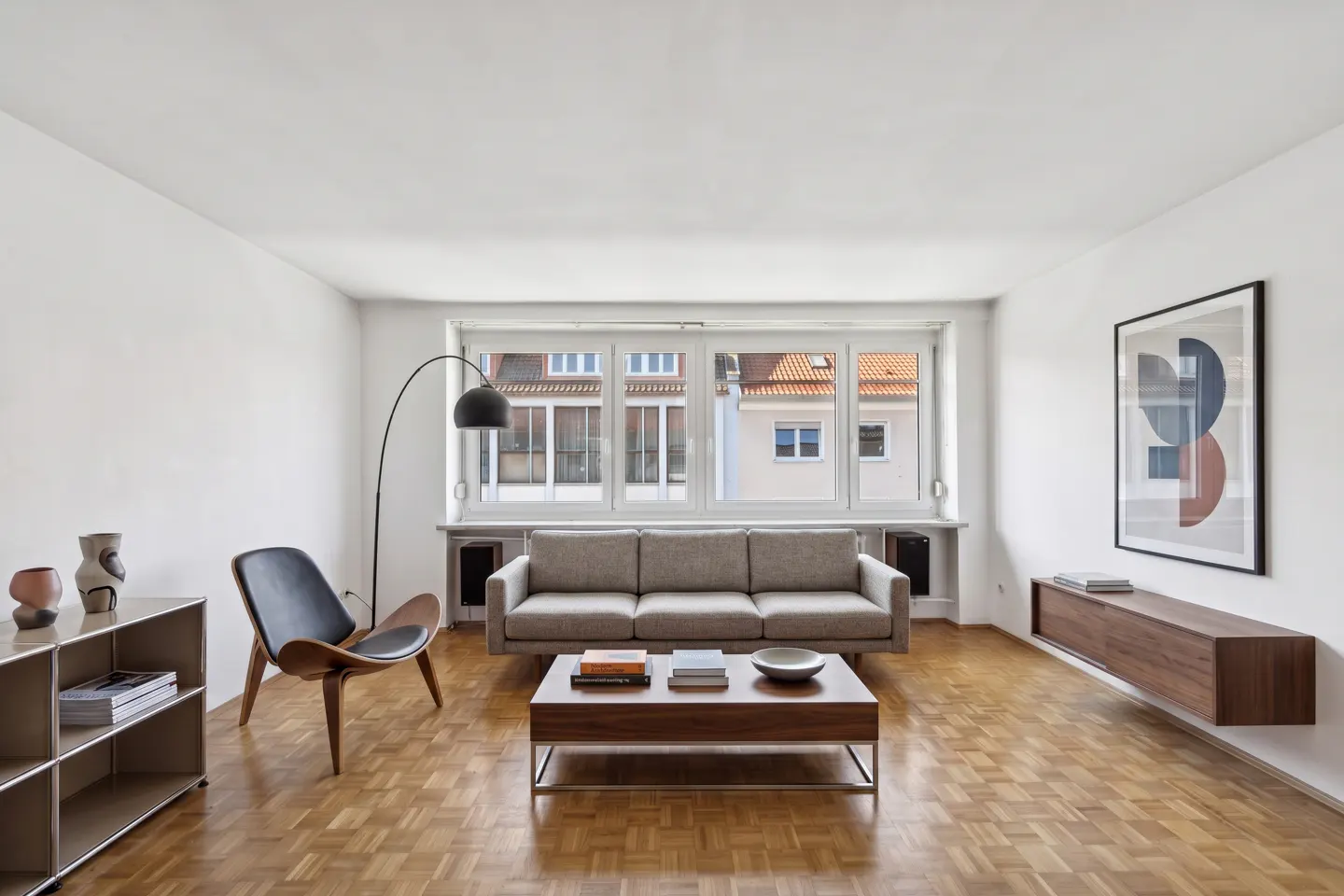 Bright living room with parquet floors, white walls, and a large window. A gray sofa faces a wooden coffee table. A black floor lamp and chair are on the left.