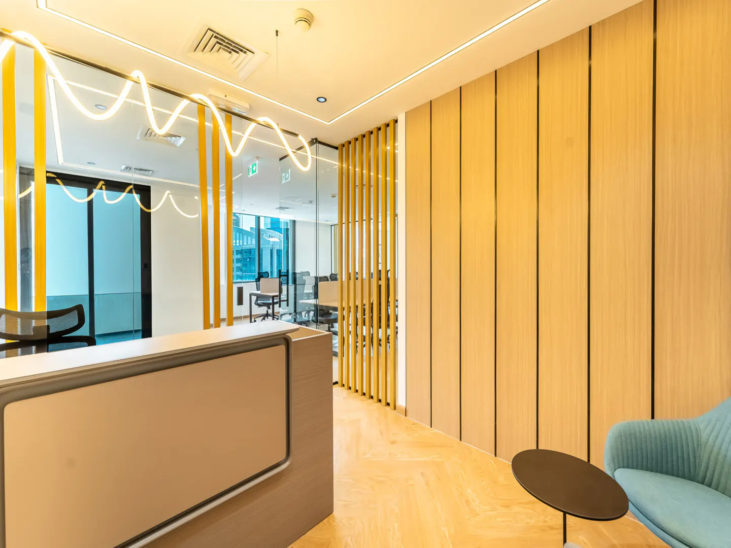 Office reception area with a beige desk, light wood walls, and a blue chair next to a small black table. A glass-walled conference room is visible in the background.