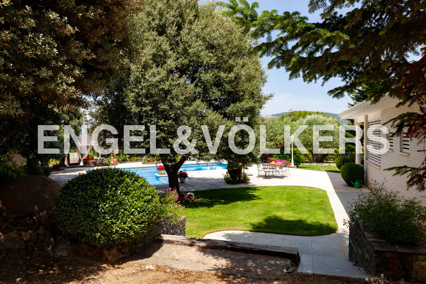 Lush green lawn with a pool and patio furniture, framed by trees and a white brick house. Engel & Völkers logo overlay.
