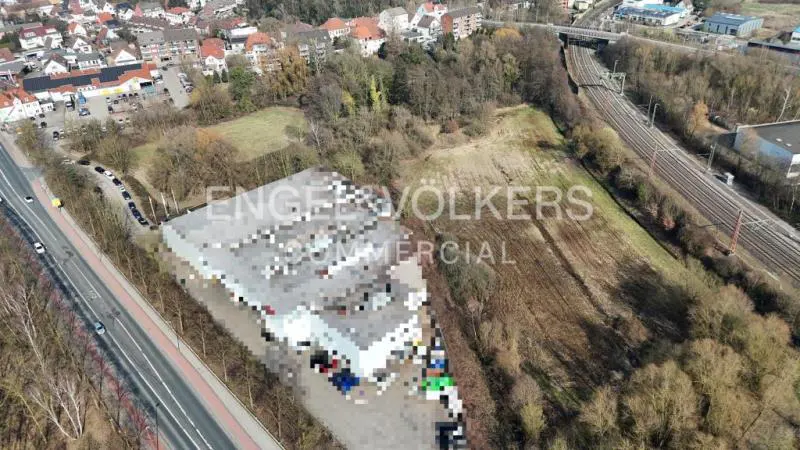 Aerial view of a gray commercial building next to a road and a field with trees. A train track is visible in the background.