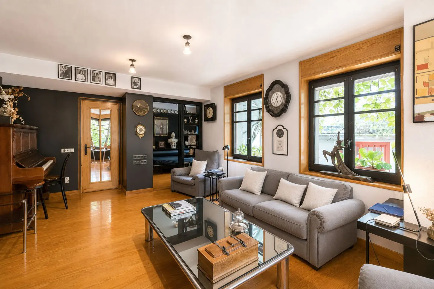 Living room with hardwood floors, gray sofas, and a glass coffee table. A piano sits against a dark wall, and windows offer a view of greenery.